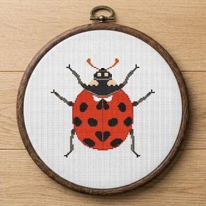 May include: A cross-stitch embroidery of a ladybug in a wooden hoop. The ladybug is red with black spots and a black head. The hoop has a metal clasp for hanging. The background is a light brown wood.