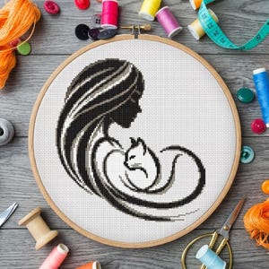 Könnte beinhalten: Ein Kreuzstich-Stickrahmen mit einer schwarz-weißen Silhouette einer Frau und einer Katze. Das Design befindet sich auf weißem Stoff, der von einem Holzrahmen umgeben ist. Nähutensilien wie Garnrollen und Scheren umgeben den Rahmen.