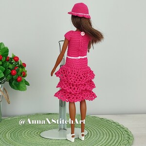 May include: A doll wearing a handmade, hot pink crochet outfit, including a dress with tiered ruffles, a short-sleeved top, and a matching hat. The outfit has white trim and is accessorized with white shoes. The doll is standing on a metal stand.