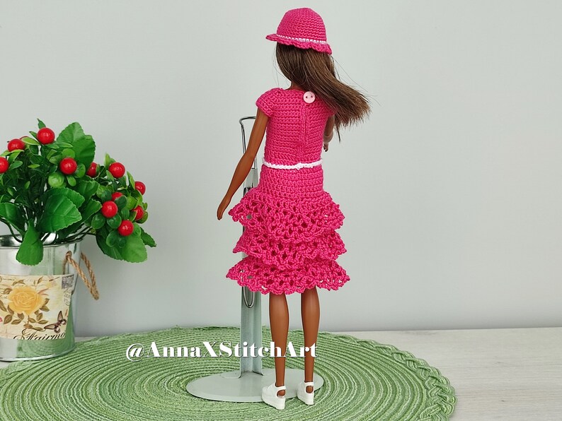 May include: A doll wearing a handmade, hot pink crochet outfit, including a dress with tiered ruffles, a short-sleeved top, and a matching hat. The outfit has white trim and is accessorized with white shoes. The doll is standing on a metal stand.