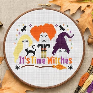 Op de afbeelding: Een borduurring met kruissteek met een Halloween-ontwerp. Het ontwerp bevat drie heksenfiguren, een zwarte kat, vleermuizen en de tekst "It's Time Witches". Het kleurenpalet omvat oranje, paars, geel en zwart.