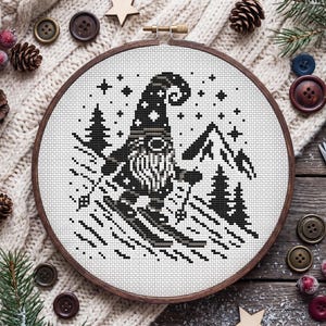 Può includere: Un telaio da ricamo circolare in legno contenente un motivo a punto croce di uno gnomo che scia su una montagna. Il disegno è realizzato con filo nero su tessuto bianco, raffigurante uno gnomo con un cappello alto, una barba e degli sci.