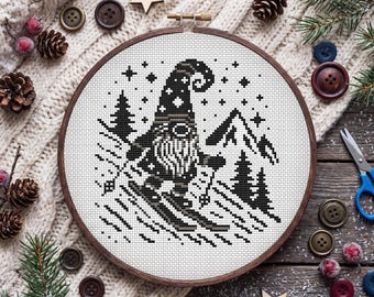 May include: A circular wooden embroidery hoop containing a cross-stitch pattern of a gnome skiing down a mountain. The design is rendered in black thread on white fabric, featuring a gnome with a tall hat, a beard, and skis, surrounded by pine trees and stars.