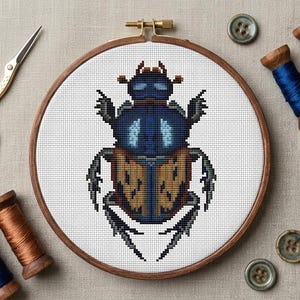 Puede incluir: Un proyecto de punto de cruz terminado de un escarabajo, exhibido en un bastidor de bordado de madera. El escarabajo es principalmente azul y naranja, con alas y cuerpo pixelados detallados. Suministros de artesanía adicionales, como tijeras, carretes de hilo y botones.
