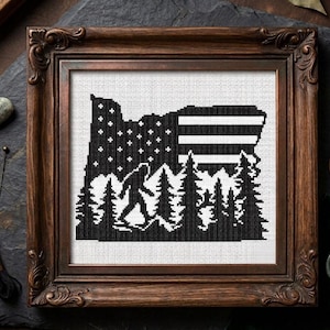 May include: A framed cross-stitch featuring the outline of Oregon, with a black and white design. The design incorporates a starry sky, stripes, trees, and a silhouette of Bigfoot. The frame is a dark brown, ornate wood.