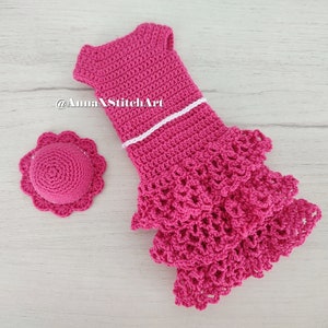May include: A handmade, hot pink crochet dress with a matching hat. The dress features a white trim and a tiered, ruffled skirt. The hat has a scalloped brim. The items are displayed on a light wood-grain surface.