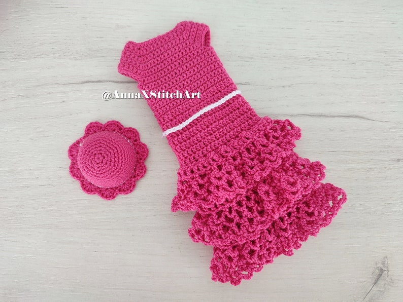 May include: A handmade, hot pink crochet dress with a matching hat. The dress features a white trim and a tiered, ruffled skirt. The hat has a scalloped brim. The items are displayed on a light wood-grain surface.