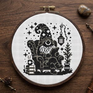 May include: A cross-stitch embroidery in a circular wooden hoop featuring a black and white design of a gnome wearing a star-patterned hat, sitting in an armchair reading a book, with a hanging lantern, a small potted plant, and a stack of books.