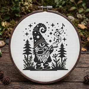 Könnte beinhalten: Ein runder Stickrahmen aus Holz mit einem Kreuzstichmuster auf weißem Stoff. Das Motiv zeigt einen schwarz-grauen Zwerg mit einer hohen Mütze, der in einer Waldlandschaft mit Tannen, Sternen und darüber schwebenden Musiknoten steht.