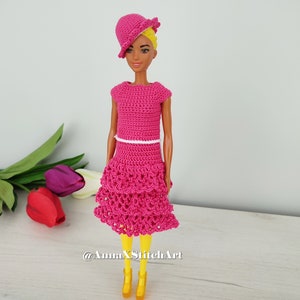 May include: A doll wearing a handmade, bright pink crochet dress with a matching hat. The dress has a tiered skirt and a white belt. The doll is also wearing yellow shoes. The background includes colorful tulips.