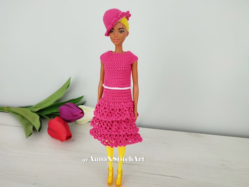 May include: A doll wearing a handmade, bright pink crochet dress with a matching hat. The dress has a tiered skirt and a white belt. The doll is also wearing yellow shoes. The background includes colorful tulips.