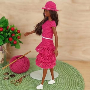 May include: A doll wearing a handmade, bright pink crochet outfit, including a hat, top, and tiered skirt. The doll is standing on a green mat, with a ball of pink yarn, a crochet hook, and a pair of scissors nearby. The outfit has a white trim.