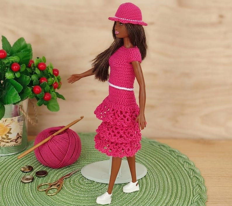 May include: A doll wearing a handmade, bright pink crochet outfit, including a hat, top, and tiered skirt. The doll is standing on a green mat, with a ball of pink yarn, a crochet hook, and a pair of scissors nearby. The outfit has a white trim.