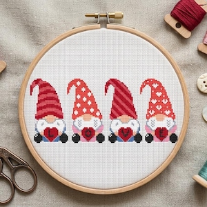 May include: A cross-stitch embroidery in a wooden hoop features four gnomes with red and white hats, each holding a heart with the word "LOVE." The gnomes have white beards and black shoes. The background is white fabric.