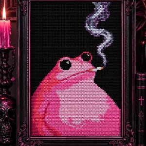 May include: A cross-stitch embroidery in a black ornate frame featuring a bright pink frog with large black eyes. The frog is depicted with a cigarette in its mouth, with a trail of grey and white smoke rising upwards against a solid black background.