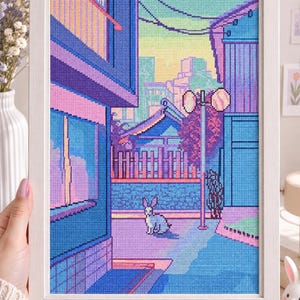 Op de afbeelding: Een ingelijst kruissteek borduurwerk met een levendig stedelijk straatbeeld bij zonsondergang. Het kunstwerk toont een klein wit konijn in het midden van een blauw en paars steegje, omringd door gebouwen, een lantaarnpaal en een verre stad.