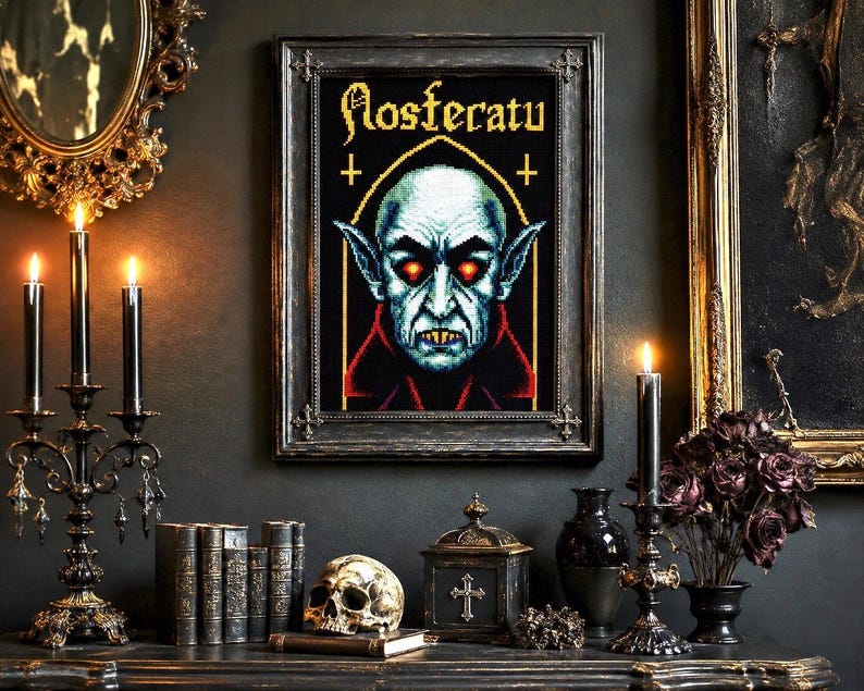 May include: A framed cross-stitch art piece featuring a pixelated portrait of the vampire Nosferatu with glowing red eyes, sharp teeth, and pointed ears. The text Nosferatu is stitched in a gothic font above the figure on a black background.