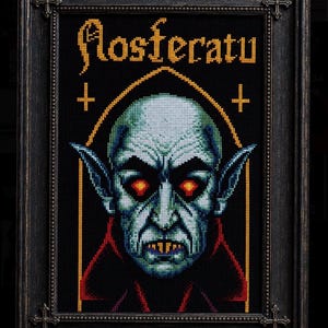 May include: A framed cross-stitch artwork featuring a pale, pointed-eared vampire with glowing red eyes and sharp teeth. The text 'Nosferatu' is stitched in yellow above the figure, flanked by two small crosses within a gold-lined arch on a black background.