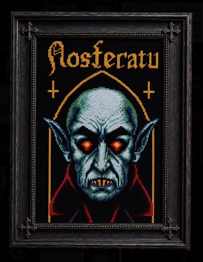May include: A framed cross-stitch artwork featuring a pale, pointed-eared vampire with glowing red eyes and sharp teeth. The text 'Nosferatu' is stitched in yellow above the figure, flanked by two small crosses within a gold-lined arch on a black background.