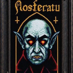 May include: A framed cross-stitch embroidery featuring a pixelated portrait of Nosferatu with pale blue skin, glowing red eyes, and yellow fangs. The text Nosferatu is stitched in gold above the figure, flanked by two small gold crosses in a black frame.
