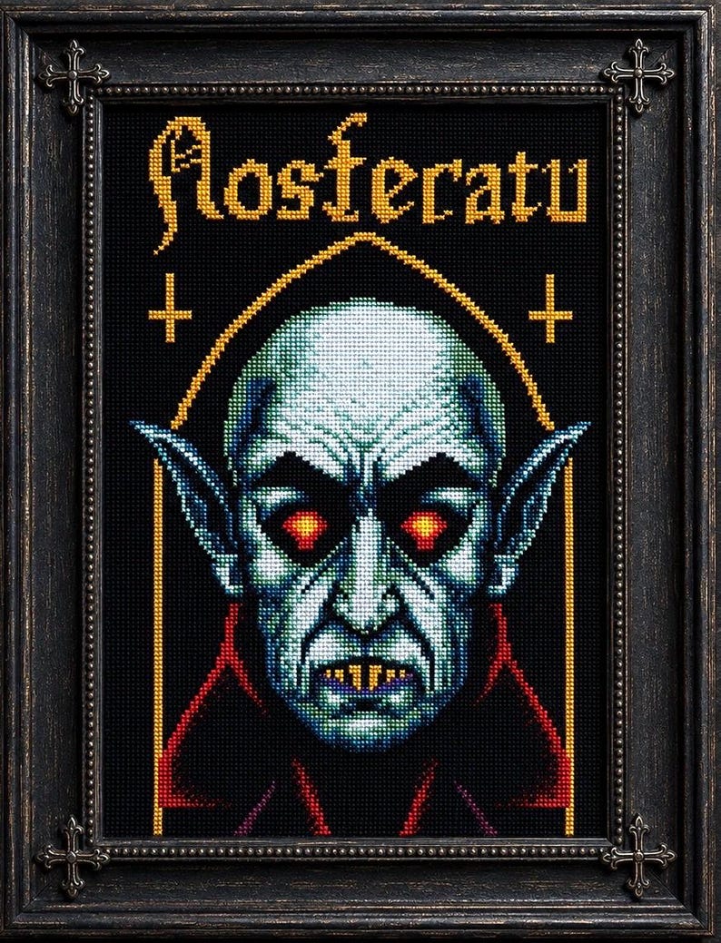 May include: A framed cross-stitch embroidery featuring a pixelated portrait of Nosferatu with pale blue skin, glowing red eyes, and yellow fangs. The text Nosferatu is stitched in gold above the figure, flanked by two small gold crosses in a black frame.