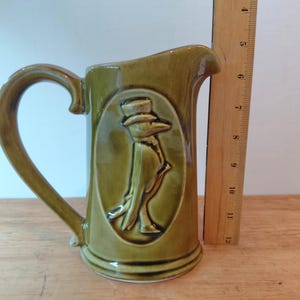 May include: An olive green ceramic pitcher featuring a raised relief design of a bird wearing a top hat within an oval frame. A wooden ruler placed vertically next to the pitcher indicates a height of approximately 6.5 inches for the vessel.