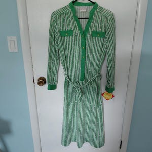May include: A knee-length dress featuring a green and white vertical striped pattern, a mandarin collar, a button-down front placket, long sleeves with cuffs, two chest pockets with flaps, and a matching fabric tie belt at the waist.