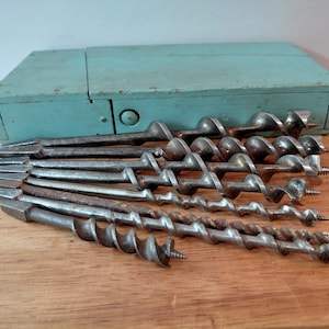 May include: A set of seven metal auger drill bits of varying diameters arranged in a fan shape, featuring spiral flutes and pointed threaded tips. The bits are positioned in front of a rectangular, light blue painted wooden storage box.