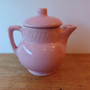 May include: A small pink ceramic teapot with a lid and a handle. The teapot has a ribbed design around the middle.