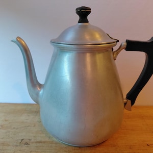 May include: A vintage aluminum coffee pot with a black handle and lid knob. The pot has a curved spout and a rounded body. The handle is black and the lid knob is black with a gold accent. The pot is sitting on a wooden surface.