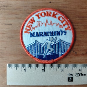 May include: A circular patch with an orange border and the words "NEW YORK CITY" in orange. The patch features a skyline and the text "MARATHON '79". A runner is depicted on a bridge in blue and white. The patch is on a wooden surface.
