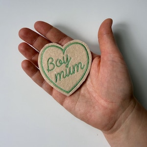Op de afbeelding: Een beige hartvormige patch met groene geborduurde tekst "Boy mum". De patch wordt in een hand gehouden, tegen een effen witte achtergrond.