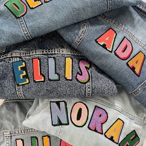 Puede incluir: Varias chaquetas vaqueras superpuestas, cada una bordada con un nombre diferente en colores vivos. Los nombres incluyen "DEXTER", "ADA", "ELLIS", "NORA" y "LILA". Los nombres están en varios colores, con contornos negros.
