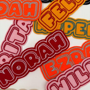 May include: A selection of felt name patches in vibrant colours like orange, pink, red, and yellow. Each patch displays a different name in a bold, outlined font with a 3D effect. These patches are suitable for customising clothing or accessories.