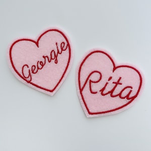 May include: Two pink felt heart-shaped patches with red embroidered outlines. The left patch says "George" and the right patch says "Rita".