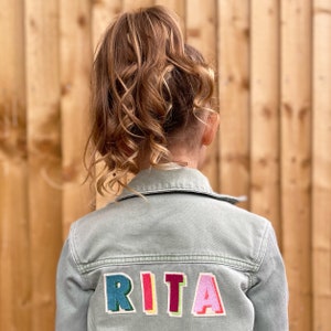 May include: A pale green denim jacket with the name "RITA" embroidered on the back in colourful, block letters. The jacket is worn by a person with light brown hair in a ponytail. The background is a wooden fence.