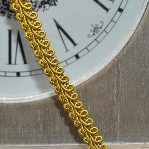 May include: A spool of gold-colored decorative trim sits next to a section of the trim draped in front of a vintage clock. The trim has a looped design and is likely used for embellishing clothing or crafts.