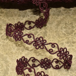 May include: A spool of burgundy decorative trim with an intricate, looped design. The trim is unwound, revealing its detailed pattern. The background features a vintage-style floral pattern.