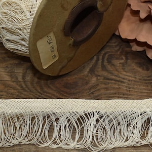 May include: A roll of cream-colored, open-weave trim with a looped fringe. The trim is displayed on a wooden spool with a label that reads "034 PER YD". A large, dried flower and a wooden surface complete the scene.
