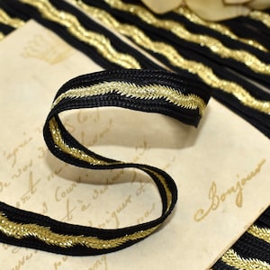 May include: Black ribbon with gold metallic trim. The ribbon is woven with a zig-zag pattern of gold thread. The ribbon is curled on a vintage paper background. The ribbon is likely used for sewing, crafting, or embellishing.