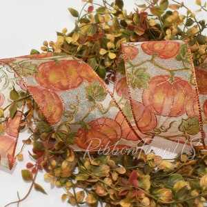 May include: A decorative ribbon with a pumpkin design. The ribbon features orange pumpkins with green leaves on a light background. The ribbon is placed on top of a bed of green and red foliage. The text "RibbonTownUS" is visible on the ribbon.