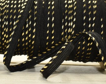 May include: A spool of black elastic ribbon with a gold metallic stripe pattern. The ribbon is tightly wound on a cardboard spool, with a length of ribbon extending out. The ribbon is on a light wood surface.