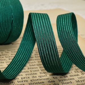 May include: A spool of dark green elastic ribbon with a ribbed texture. The ribbon is unrolled, revealing its flexibility and sheen. The background features a vintage paper with faded text.