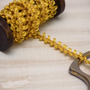 May include: A spool of yellow decorative trim with a textured, looped design. The trim is partially unraveled, revealing a gold metallic thread. A pair of scissors is in the foreground. The spool is made of dark wood.
