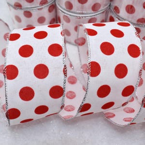 May include: White ribbon with large red polka dots. The ribbon has a silver metallic edge. Several rolls of the same ribbon are visible in the background, creating a festive and decorative theme.