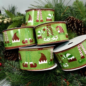 May include: Several rolls of green Christmas ribbon with an elf theme. The ribbon features elf hats, legs, and shoes in red, white, and green. The ribbon is decorated with a red border and is displayed on a bed of pine needles and pine cones.