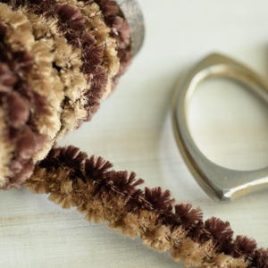 May include: Close-up of a decorative trim with a textured, fuzzy surface in shades of brown and tan. A wooden element and a silver-colored metal ring are also visible, suggesting a craft or design element.