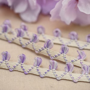 May include: Close-up of decorative trim with a cream-colored base and a repeating pattern of purple tufts. The trim is accented with a purple zigzag stitch. Soft, blurred purple flowers are in the background, suggesting a craft or sewing theme.