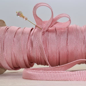 May include: A spool of pink ribbon with a woven texture, partially unraveled. A gold-colored pin with a decorative, multi-faceted bead is inserted into the ribbon. The ribbon is on a wooden spool.