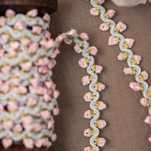 May include: A spool of pink and green ribbon with a light blue wavy trim. The ribbon is decorated with small pink flower-like accents. The ribbon is on a wooden spool, with some ribbon unspooled and laid out.